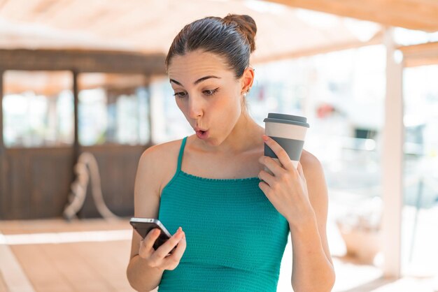 Foto mujer bonita joven al aire libre usando un teléfono móvil y sosteniendo un café con expresión sorprendida
