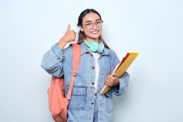 Mujer bonita joven alegre que lleva una mochila que sostiene los libros que hacen los pulgares para arriba aislados en el fondo blanco