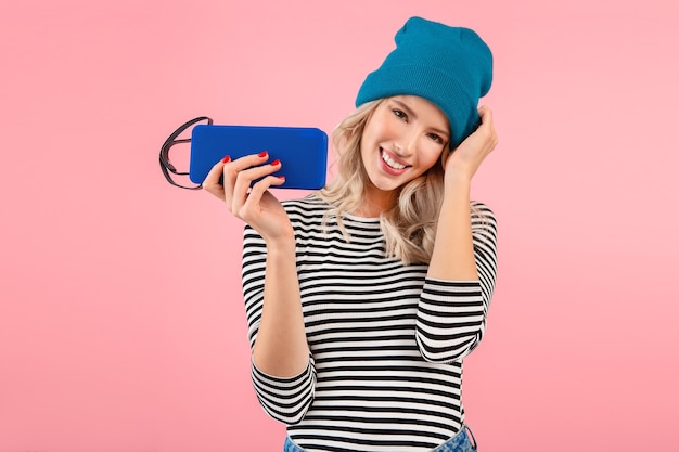 Mujer bonita joven con altavoz inalámbrico escuchando música con camisa a rayas y sombrero azul sonriendo feliz estado de ánimo positivo posando sobre fondo rosa