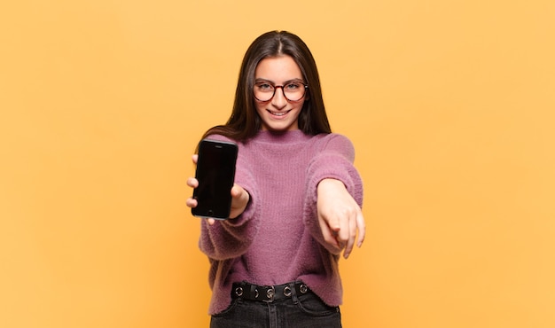 Mujer bonita joven apuntando al frente con una sonrisa satisfecha, confiada y amistosa, eligiéndote. concepto de pantalla de teléfono