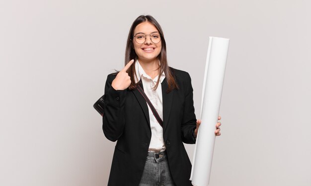 Mujer bonita joven arquitecto sonriendo con confianza apuntando a su propia sonrisa amplia, actitud positiva, relajada y satisfecha