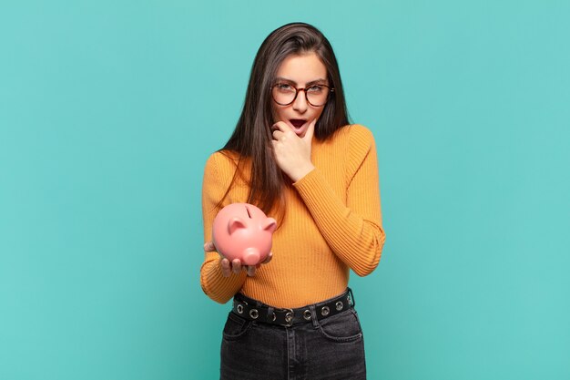 Mujer bonita joven con la boca y los ojos bien abiertos y la mano en la barbilla, sintiéndose desagradablemente conmocionada, diciendo qué o guau. concepto de alcancía