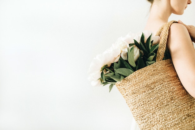 Mujer bonita joven con bolsa de paja con ramo de flores de peonía blanca sobre fondo blanco. Concepto de belleza de verano.