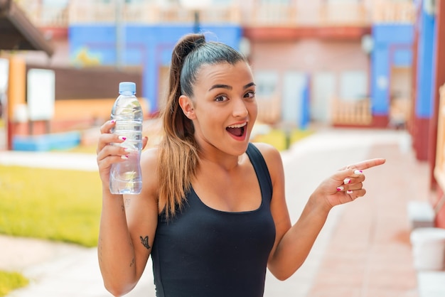 Mujer bonita joven con una botella de agua al aire libre sorprendida y señalando con el dedo hacia un lado