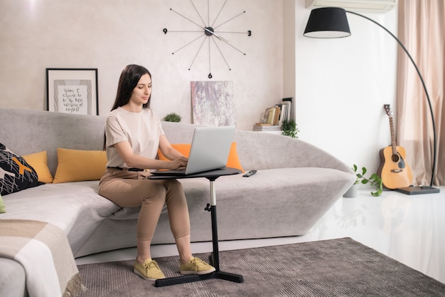 Foto mujer bonita joven con cabello largo oscuro sentado en el sofá frente a una mesa pequeña con una computadora portátil y tocar los botones del teclado en la sala de estar