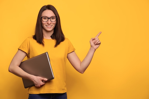 Mujer bonita joven de cabello oscuro en anteojos está sonriendo, sosteniendo la computadora portátil y apuntando