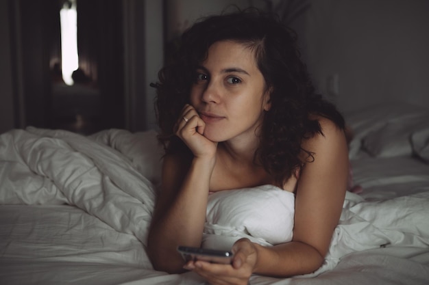 Mujer bonita joven con cabello rizado en la cama con teléfono