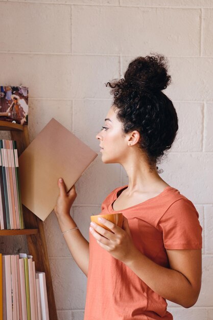 Mujer bonita joven con cabello rizado oscuro en camiseta sosteniendo una taza de café en la mano eligiendo cuidadosamente un libro de la estantería en un hogar acogedor