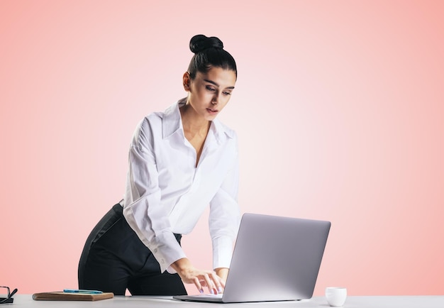 Mujer bonita joven con camisa blanca escribiendo en el teclado de un portátil moderno aislado en un primer plano de fondo de pared rosa claro abstracto