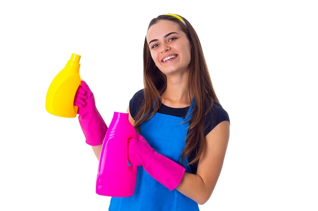 Mujer bonita joven con camiseta azul y delantal con guantes rosas sosteniendo dos detergentes en el estudio
