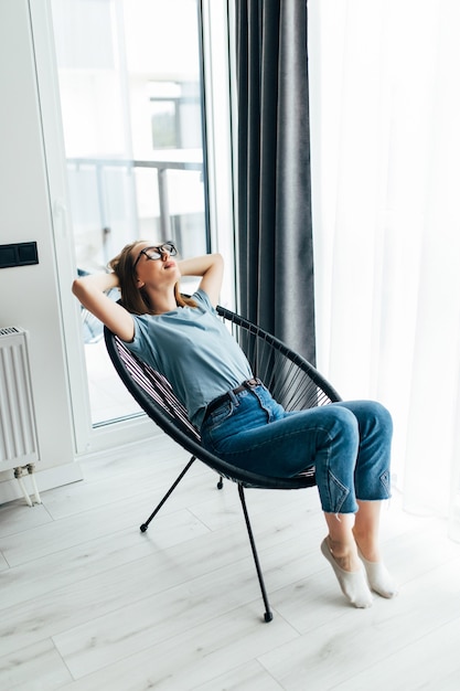 Mujer bonita joven en casa sentado en una silla moderna delante de la ventana relajándose en su sala de estar