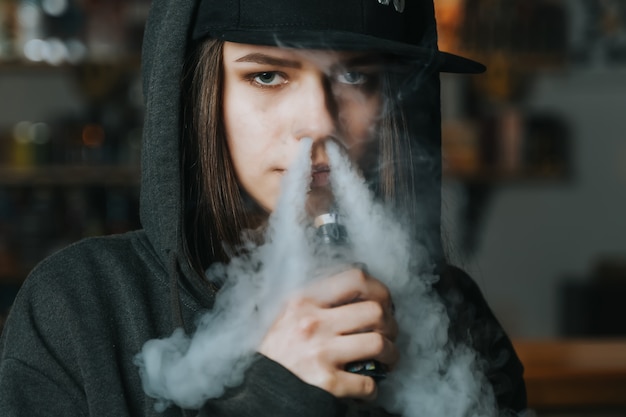 La mujer bonita joven en casquillo fuma un cigarrillo electrónico en la tienda del vape. Estilo hip-hop De cerca.