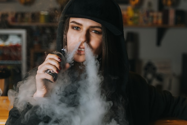 La mujer bonita joven en casquillo fuma un cigarrillo electrónico en la tienda del vape. Estilo hip-hop De cerca.