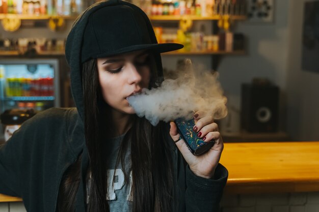 La mujer bonita joven en casquillo fuma un cigarrillo electrónico en la tienda del vape. Estilo hip-hop De cerca.