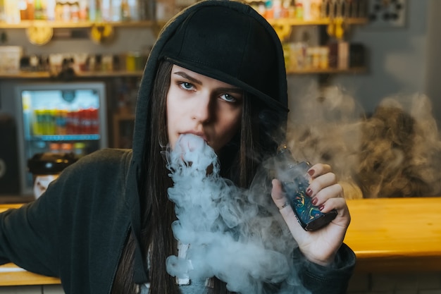 La mujer bonita joven en casquillo fuma un cigarrillo electrónico en la tienda del vape. Estilo hip-hop De cerca.