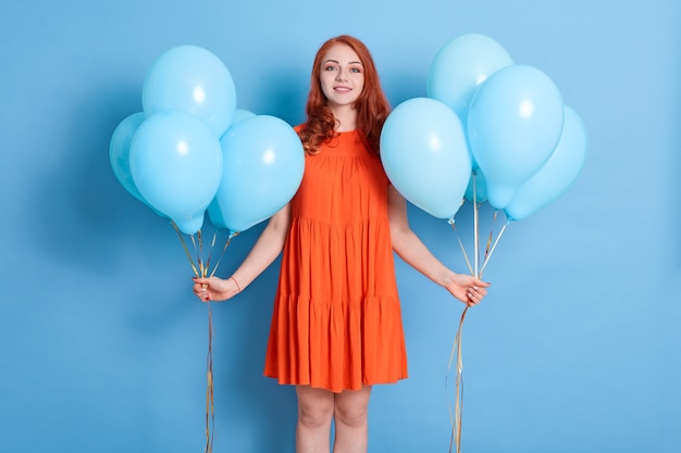 Mujer bonita joven con dos racimos de globos de pie contra la pared azul