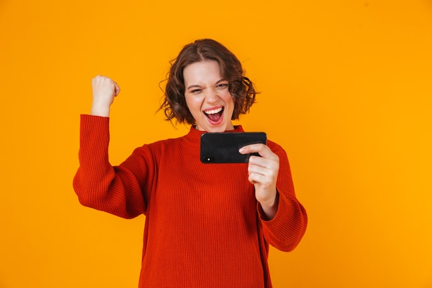 mujer bonita joven emocional posando aislada en la pared amarilla jugar juegos por teléfono móvil hacer gesto ganador.