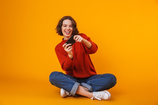 Mujer bonita joven emocional posando aislada en la pared amarilla usando juegos de teléfono móvil.