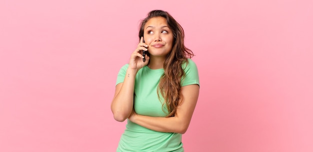 Foto mujer bonita joven encogiéndose de hombros, sintiéndose confundida e insegura y sosteniendo un teléfono inteligente