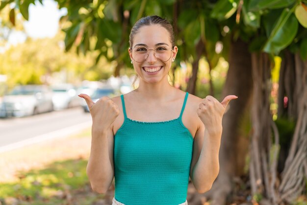 Mujer bonita joven con gafas al aire libre con gesto de pulgares hacia arriba y sonriendo