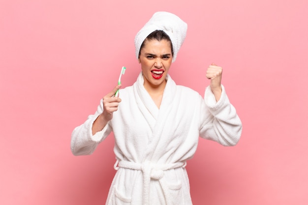 Mujer bonita joven gritando agresivamente con una expresión de enojo o con los puños cerrados celebrando el éxito. concepto de albornoz y cepillo de dientes