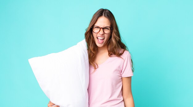 Mujer bonita joven gritando agresivamente, luciendo muy enojado vistiendo pijama y sosteniendo una almohada