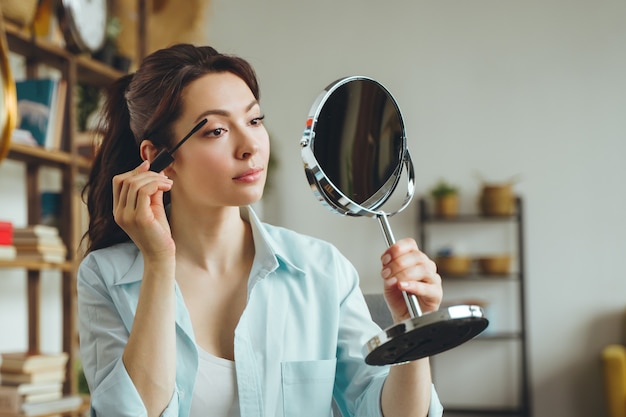 Mujer bonita joven hace maquillaje en casa mientras se mira en el espejo