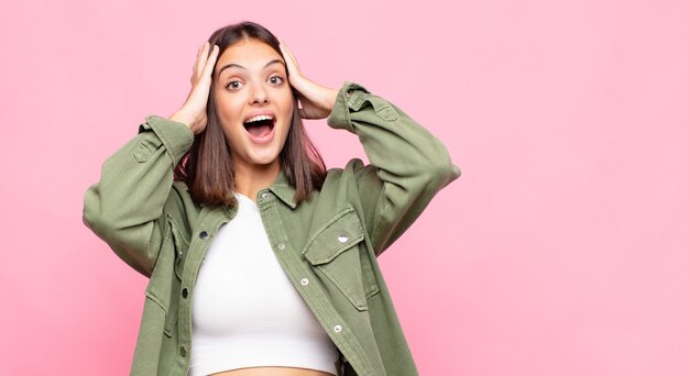 Foto mujer bonita joven levantando las manos a la cabeza, con la boca abierta, sintiéndose extremadamente afortunada, sorprendida, emocionada y feliz