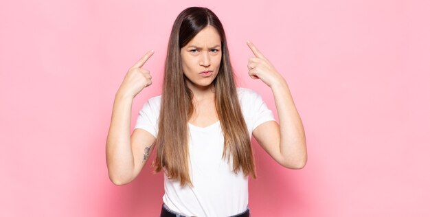 Mujer bonita joven con una mala actitud que parece orgullosa y agresiva, apuntando hacia arriba o haciendo un signo divertido con las manos