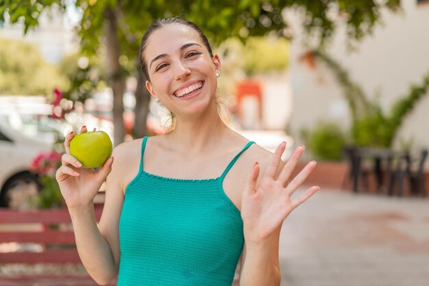 Mujer bonita joven con una manzana al aire libre saludando con la mano con expresión feliz