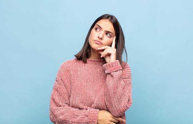 Mujer bonita joven con una mirada concentrada, preguntándose con una expresión dudosa, mirando hacia arriba y hacia un lado