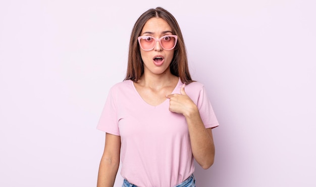 Mujer bonita joven mirando conmocionado y sorprendido con la boca abierta, apuntando a sí mismo. concepto de gafas de sol rosa