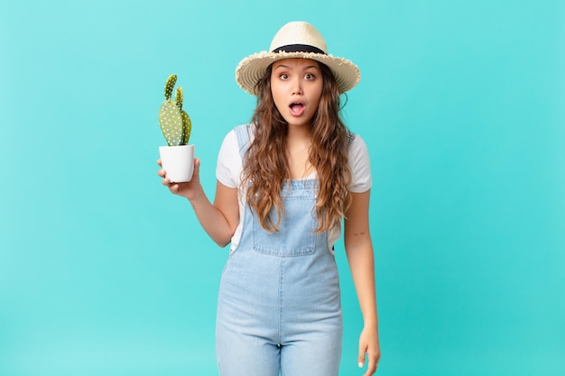 Mujer bonita joven mirando muy conmocionado o sorprendido y sosteniendo un cactus