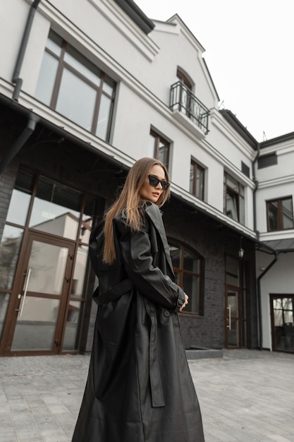 Mujer bonita joven de moda con gafas de sol vintage en elegante abrigo largo de cuero negro camina cerca de un edificio en la calle
