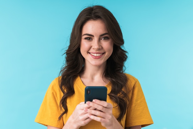 una mujer bonita joven optimista feliz posando aislada sobre la pared azul mediante teléfono móvil.
