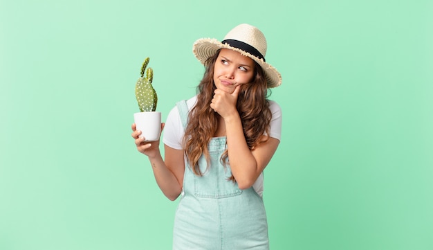 Mujer bonita joven pensando, sintiéndose dudoso y confundido y sosteniendo un cactus