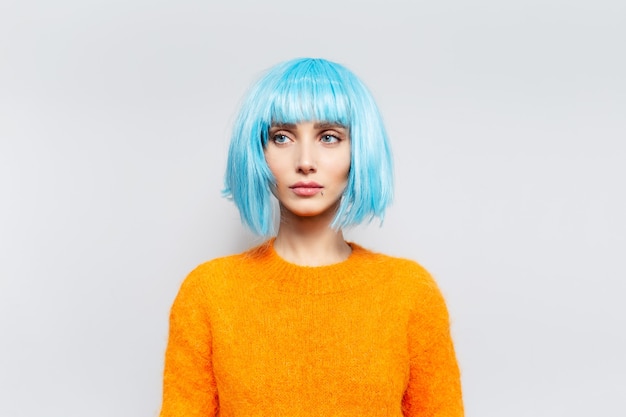 Foto mujer bonita joven pensativa con el pelo azul en suéter naranja contra la pared blanca.