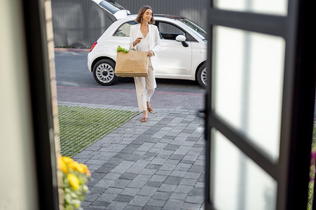 Mujer bonita joven que va a casa con una bolsa de compras llena de alimentos frescos, que llega a casa en un coche de la ciudad. Ver a través de la puerta de la casa