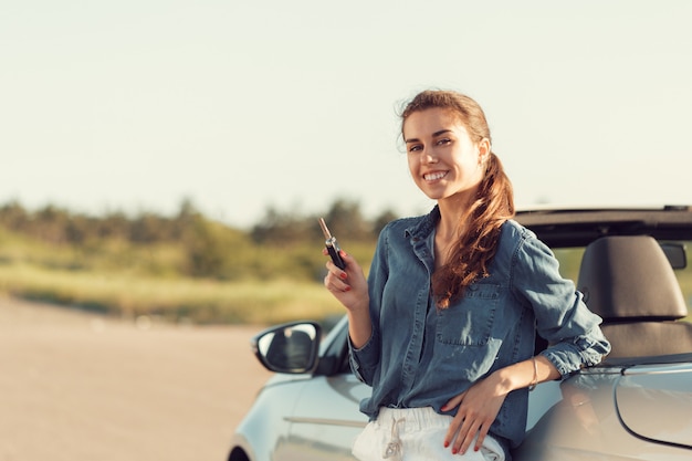 Mujer bonita joven que se coloca cerca del convertible con llaves a disposición