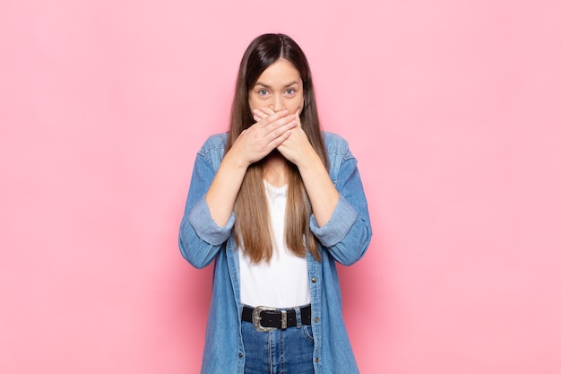 Mujer bonita joven que cubre la boca con las manos con una expresión de asombro y sorpresa, manteniendo un secreto o diciendo uy