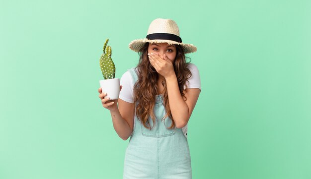 Mujer bonita joven que cubre la boca con las manos con un sorprendido y sosteniendo un cactus