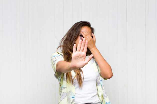 Mujer bonita joven que cubre la cara con la mano y pone la otra mano al frente para detener la cámara, rechazando fotos o imágenes