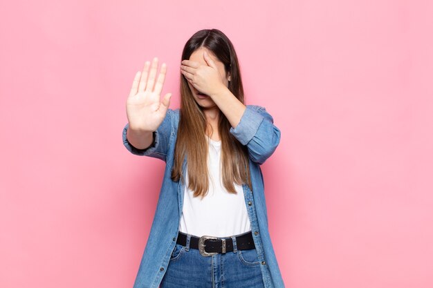 Mujer bonita joven que cubre la cara con la mano y pone la otra mano al frente para detener la cámara, rechazando fotos o imágenes