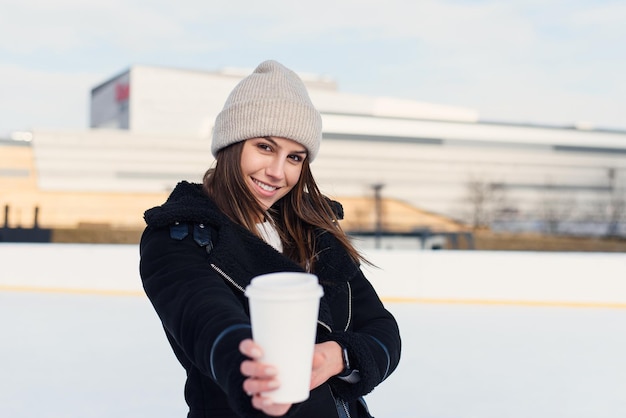 Mujer bonita joven que da ofrece una taza de café de pie en la pista de hielo