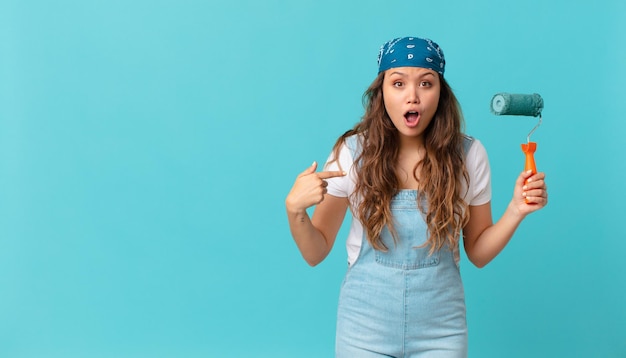 Mujer bonita joven que mira conmocionado y sorprendido con la boca abierta, apuntando a sí mismo y pintando una pared