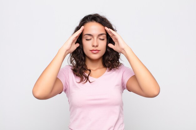 Mujer bonita joven que parece concentrada, reflexiva e inspirada, lluvia de ideas e imaginación con las manos en la frente contra la pared blanca