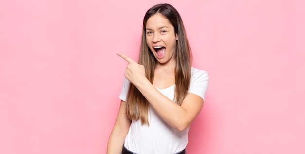 Mujer bonita joven que parece emocionada y sorprendida apuntando hacia un lado y hacia arriba para copiar el espacio