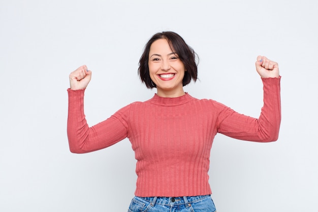 Mujer bonita joven que parece extremadamente feliz y sorprendida, celebrando el éxito, gritando y saltando contra la pared blanca