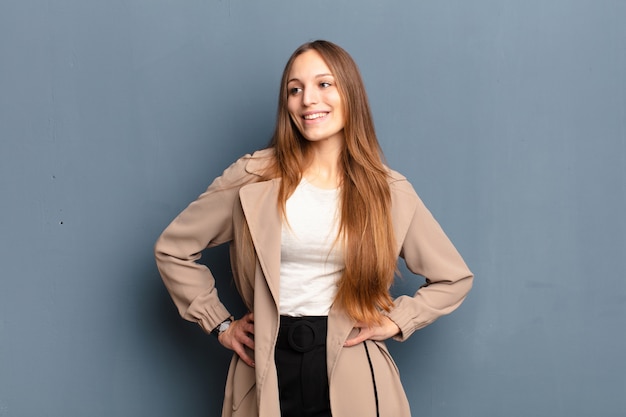 Mujer bonita joven que parece feliz, alegre y confiada, sonriendo con orgullo y mirando al lado con ambas manos en las caderas contra el fondo gris