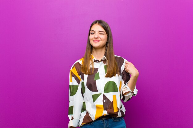 Mujer bonita joven que parece feliz y amigable, sonriendo y guiñándole un ojo con una actitud positiva sobre fondo púrpura